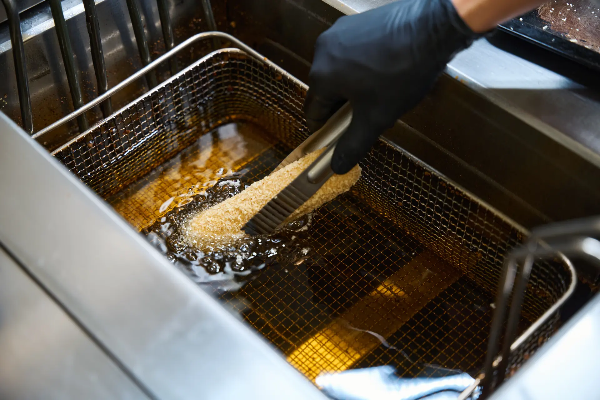 A hand in a black glove deep-fries breaded food using metal tongs in a stainless steel fryer filled with oil.
